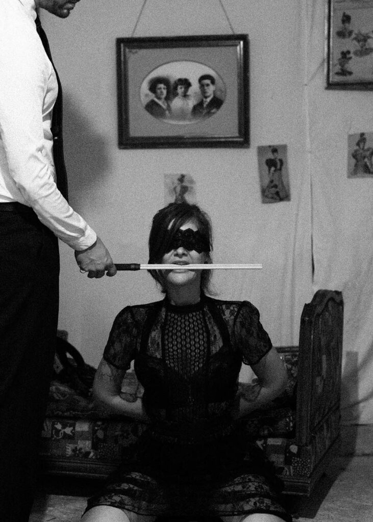 le bras d'un homme portant une badine tenue dans la bouche de sa compagne; a genoux, les yeux bandés, un rituel de soumission. Esthétique du désir, l'Amour et la violence x Fessée artisanale