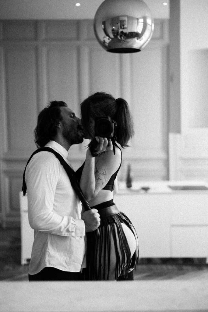 photo d'un couple s'embrassant devant le miroir, elle porte la jupe a franges de la fessée artisanale et lui en chemine blanche un martinet de la fessée artisanale en main, les franges sur l'épaule. Esthétique du désir, l'Amour et la violence x Fessée artisanale