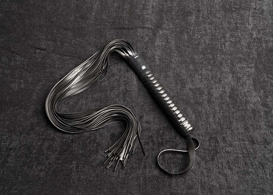 Photo montrant entièrement un martinet modèle Drash 65cm de lanières et tressage ozier en caoutchouc noir de la marque Fessée Artisanale sur fond sombre.