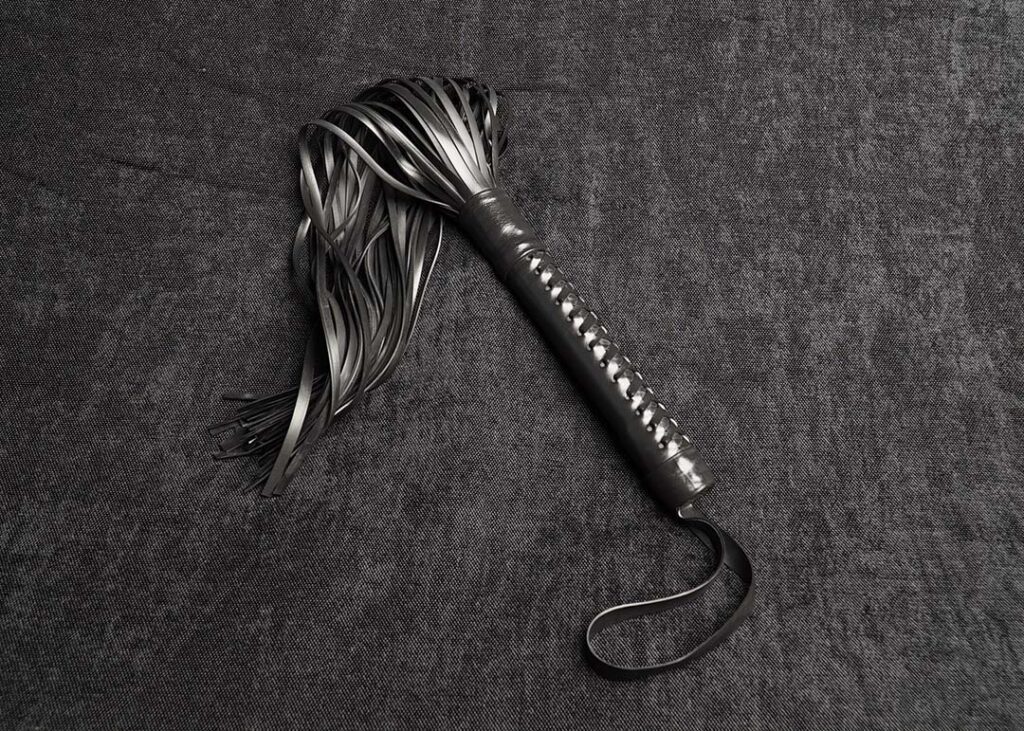 Photo montrant entièrement un martinet modèle Epona 35cm de lanières et tressage Corset en caoutchouc noir de la marque Fessée Artisanale sur fond sombre.