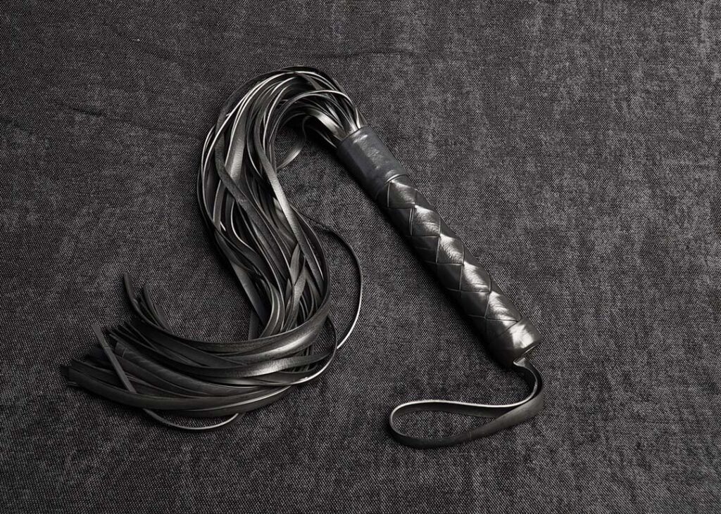 Photo montrant entièrement un martinet modèle Exalt 45cm de lanières et tressage Bonnet-turc en caoutchouc noir de la marque Fessée Artisanale sur fond sombre.