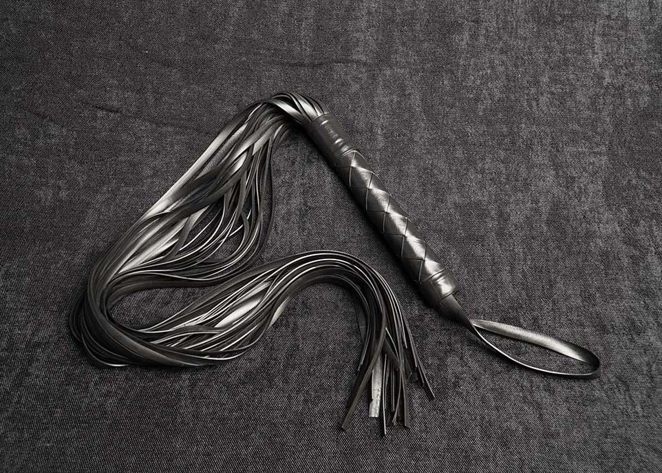 Photo montrant entièrement un martinet modèle Vega 70cm de lanièrestressage Bonnet turc en caoutchouc noir de la marque Fessée Artisanale sur fond sombre.