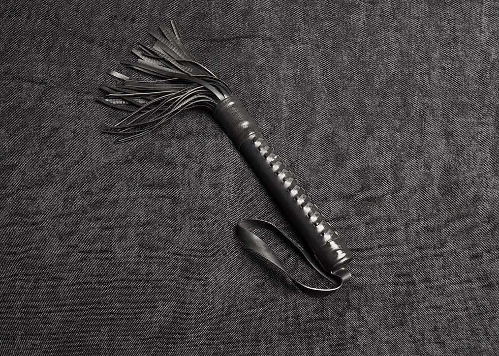 Photo montrant entièrement un martinet modèle Nebra 12cm de lanières et tressage ozier en caoutchouc noir de la marque Fessée Artisanale sur fond sombre.